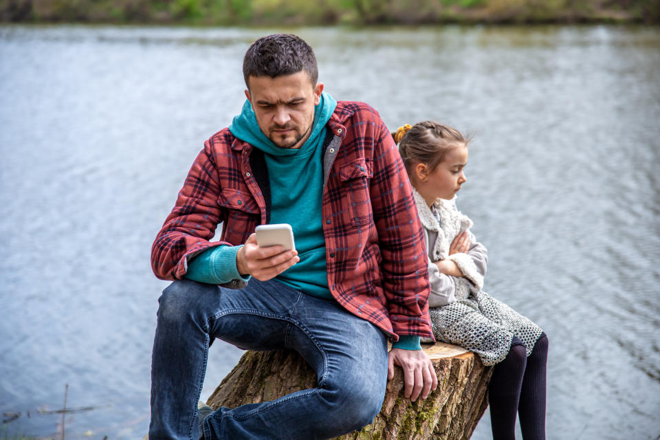 Man in a hooded jacket looks at his phone while sitting on a tree stump; a young girl in a coat stands nearby with arms crossed, both by a lake