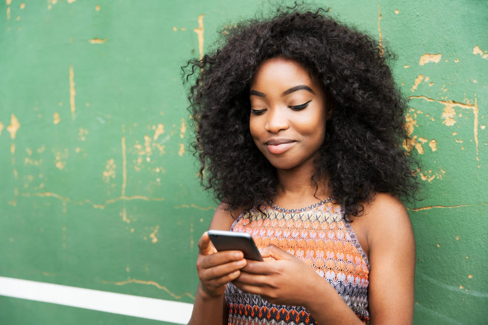 A person with curly hair smiles while looking at their phone, leaning against a textured wall