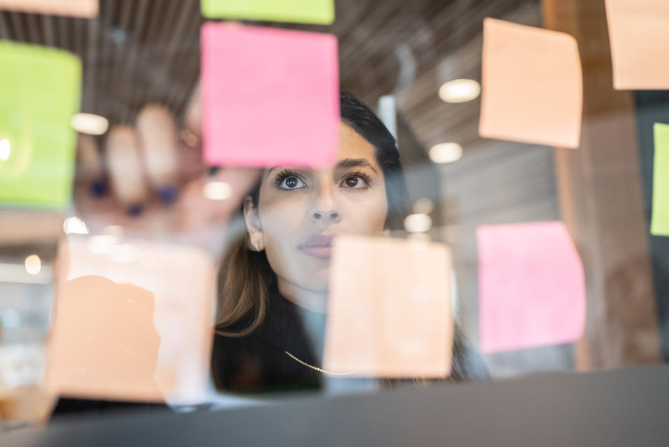 A woman is placing sticky notes on a glass wall, focusing intensely on her task. She appears to be organizing her thoughts or planning