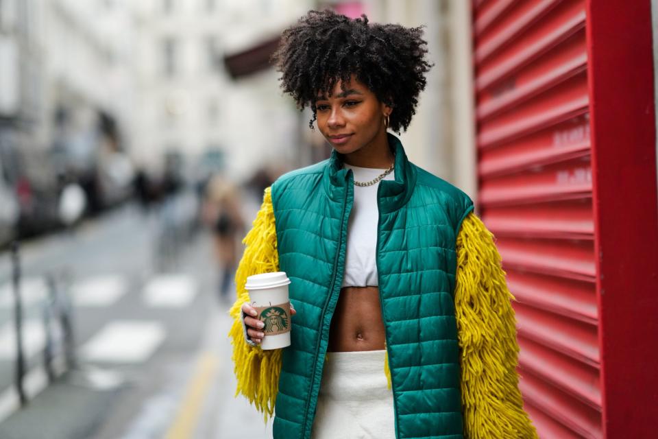 A guest wears gold large earrings, a gold large chain necklace, a white cropped t-shirt with yellow large fluffy fringes long sleeves, a dark green sleeveless puffer jacket, a high waist felt midi tube skirt, an orange suede and brown leather handbag, outside Louis Vuitton, during Paris Fashion Week January 2022 - Copyright: Berthelot/Getty Images