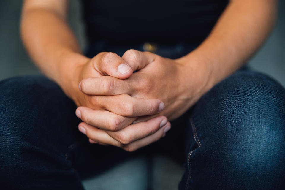 A person's hands are clasped together, resting on their lap, with the individual wearing dark jeans.