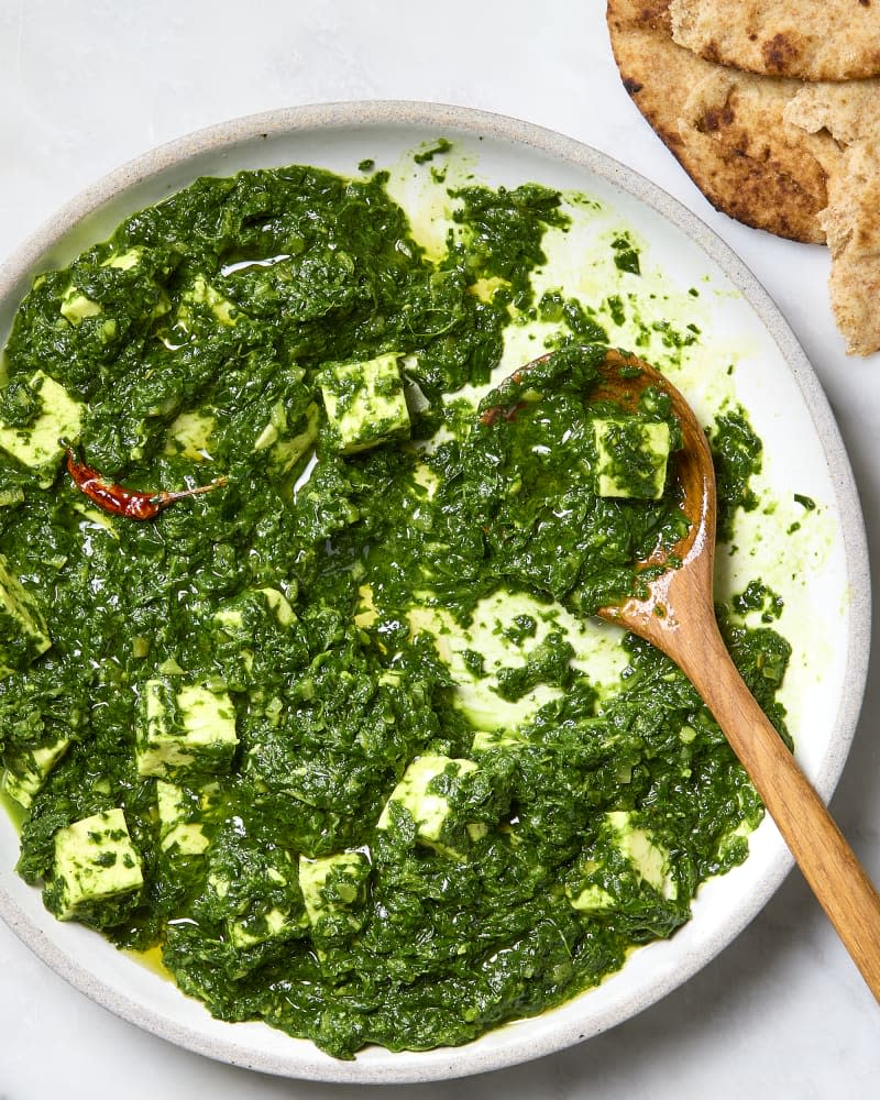 A platter of saag paneer with some food missing with a wooden serving spoon. There is whole wheat pita bread on the side.