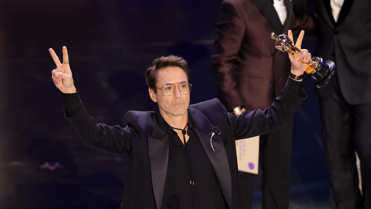 robert downey jr holds an oscar in one hand as he flashes two peace signs