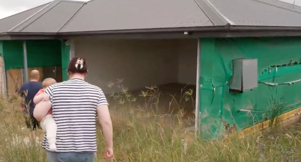 The couple walk through long grass toward the half-built home. 