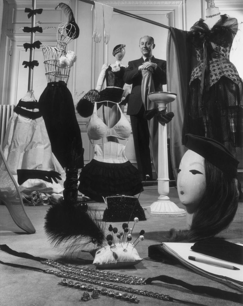 French fashion designer Christian Dior standing in a showroom with samples of his design accessories for women, including hats, hat pins, gloves, muffs, lingerie, hosiery, evening bags, and jewelry.