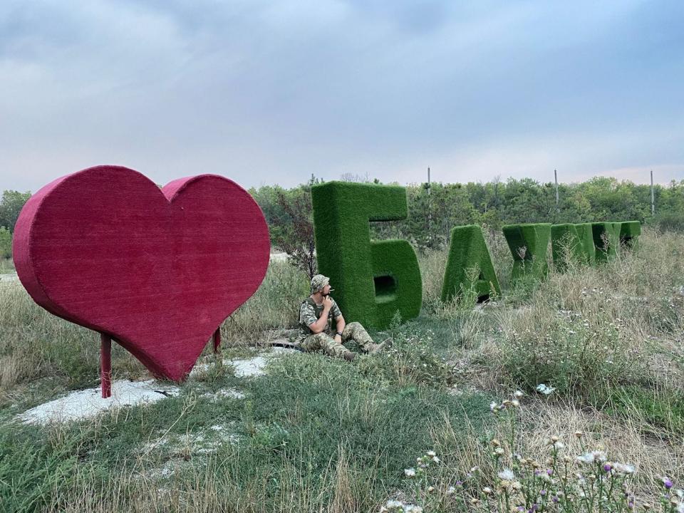 Borys Khmelevskiy, in camouflage gear, sits in grass in front of a large fake grass sign that says "<3 Bakhmut"