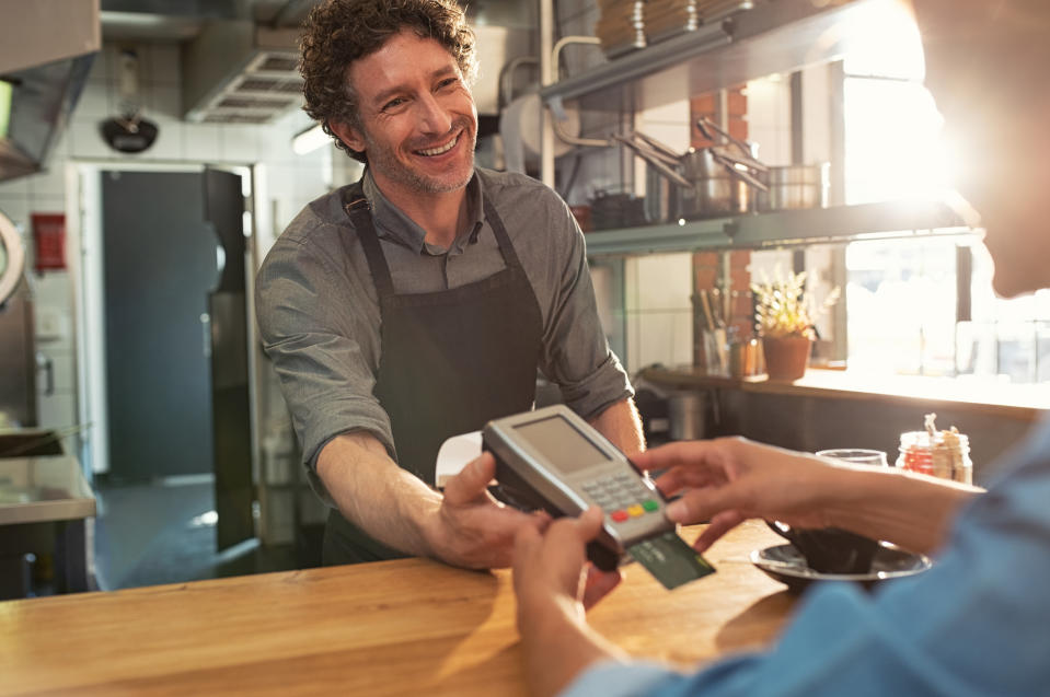 A smiling male barista holds out a card reader to a customer.