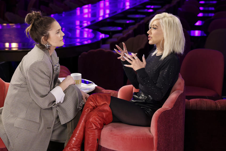 Drew Barrymore and Doja Cat engaged in a conversation while seated on a talk show set. Drew is in a plaid suit, and Doja is in a stylish outfit with red boots