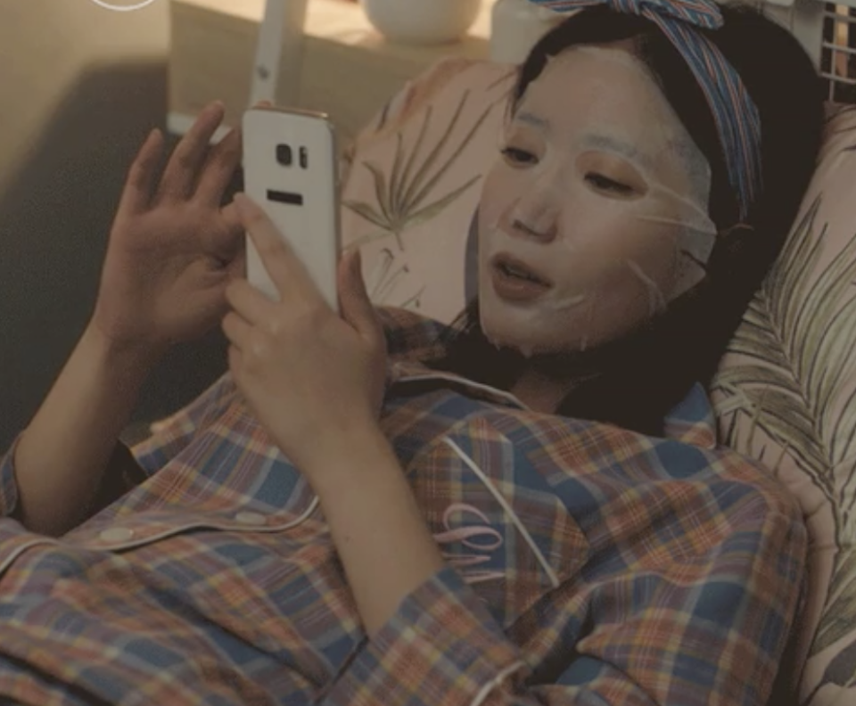 A woman is wearing a sheet mask and scrolling through her phone