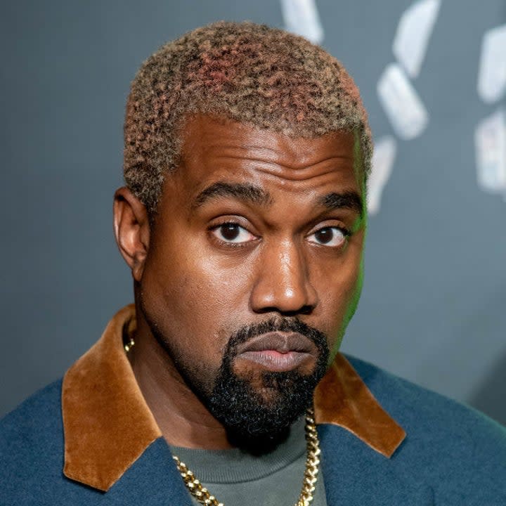 Headshot of Kanye with bleached hair and a goatee, he rocks a gold chain and blue suit with tan suede collar