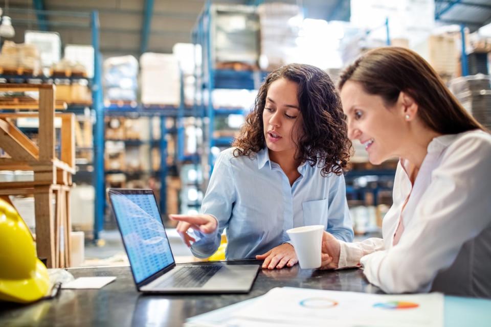 Two people sit at a table in a warehouse, discussing what is happening on a computer screen. 