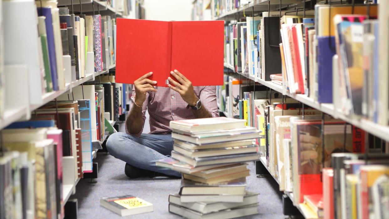  Person sat on floor in library reading red book. 