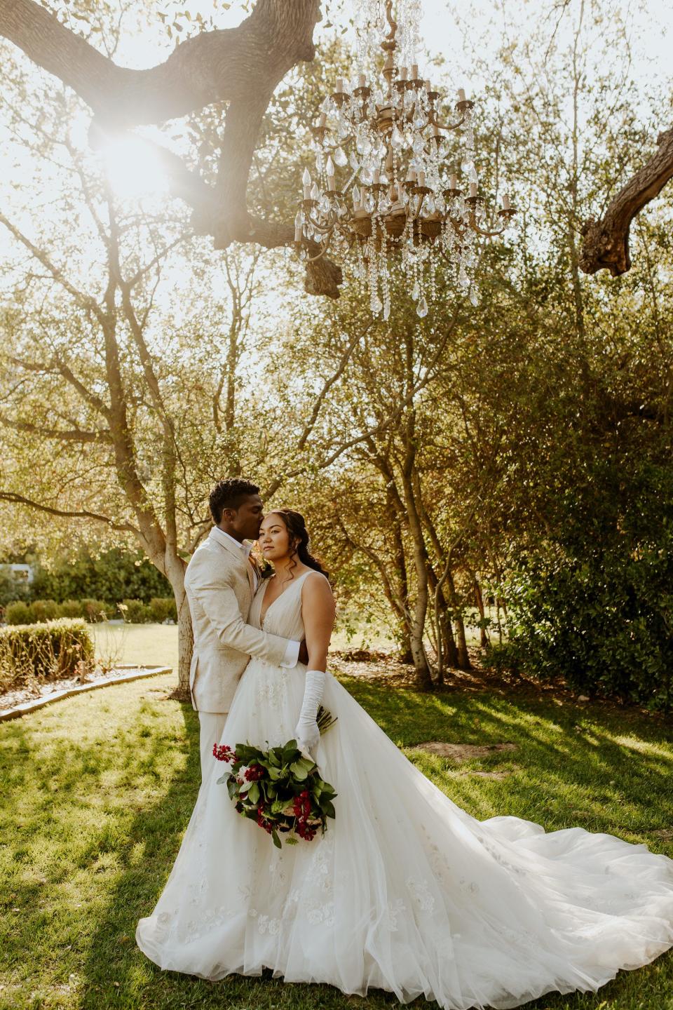 A bride and groom embrace in a field surrounded by trees.