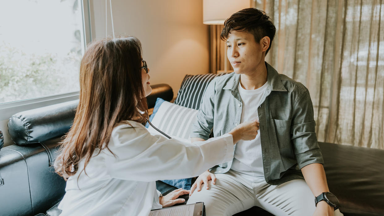  Asian female doctor using stethoscope listening the heartbeat of transgender man seated on an examination table. 