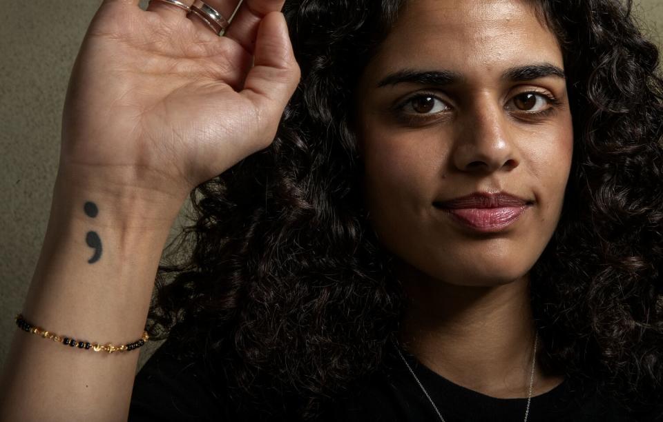 A woman with curly hair and a black semicolon tattoo on her right arm smiles for a photo.