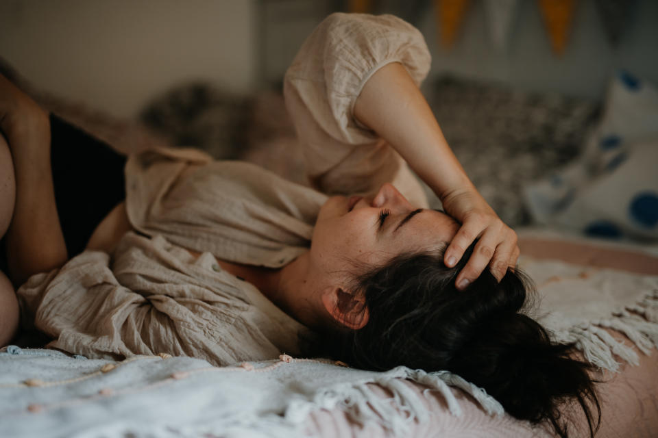 A woman lies on her back in bed, with her hand resting on her forehead, looking relaxed and thoughtful