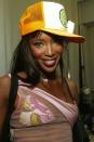 <p>Campbell serving a look backstage in 2002 with purple lipstick, matching blush, colored contacts, and a trucker hat (because of course).</p>