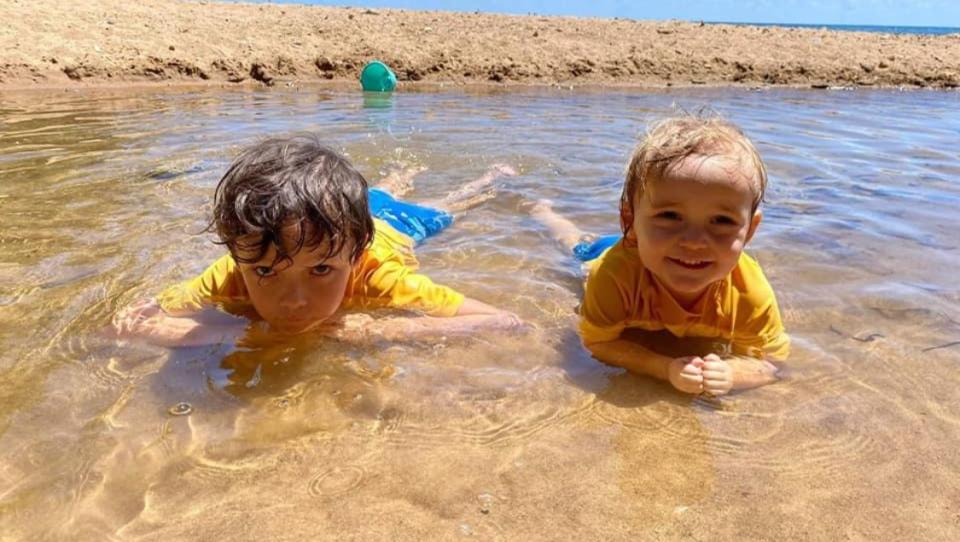 Two young boys in yellow rash shirts lying in water smiling at camera.