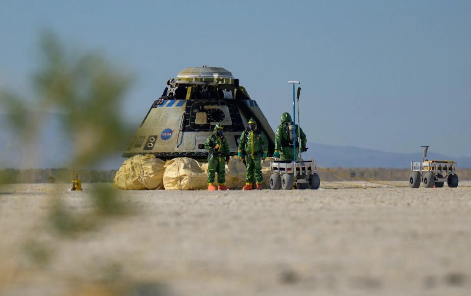 Three people in green hazmat suits stand next to a space capsule, sitting in the desert on large airbags next to two metal rover-like carts