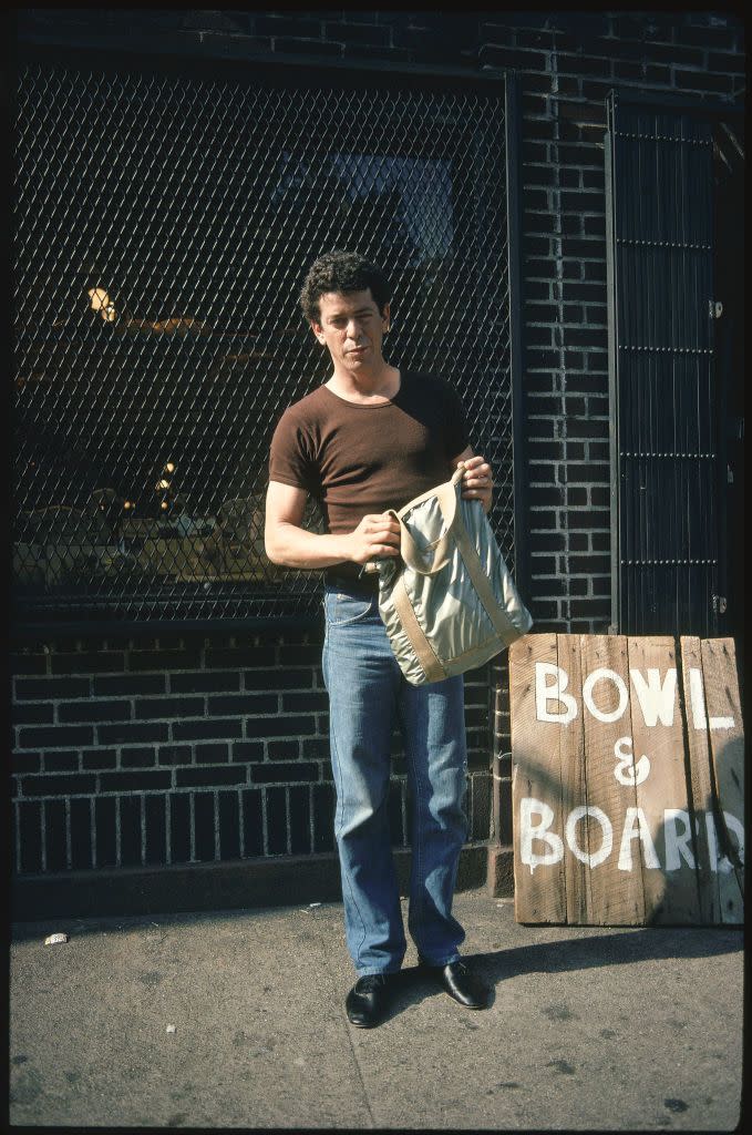 lou reed holds a bag in both hands as he stands on a sidewalk in front of brick building, he wears a brown short sleeve shirt and jeans