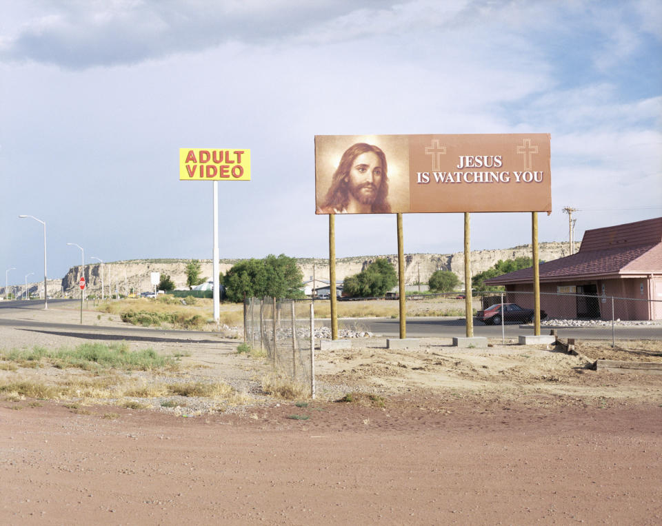 Billboard near an "Adult Video" store displays an image of Jesus with the text "JESUS IS WATCHING YOU"