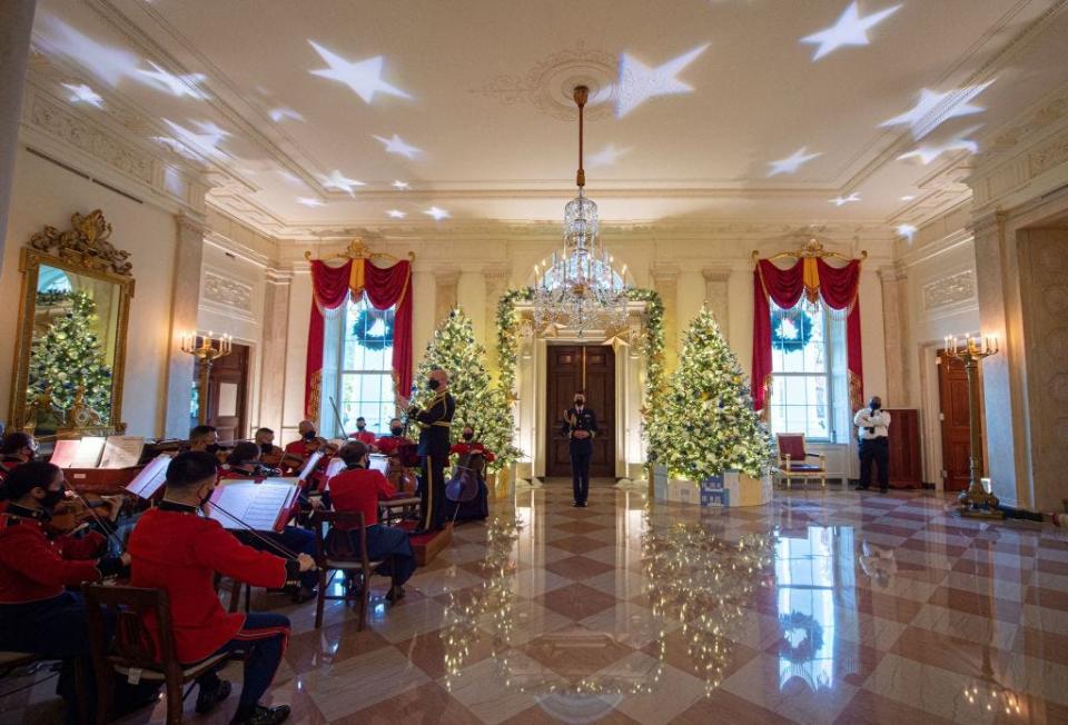 The Marine Band plays holiday music in the White House, decorated for Christmas with trees and stars projected on the ceiling.