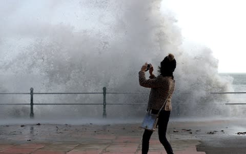  A woman take a photograph as waves crash up onto Penzance seafront - Credit: Matt Cardy/Getty