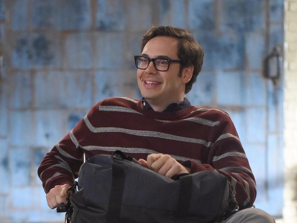 Nelson Franklin in a wheelchair wearing a maroon and gray striped shirt with a cast on his right leg on the set of "New Girl"