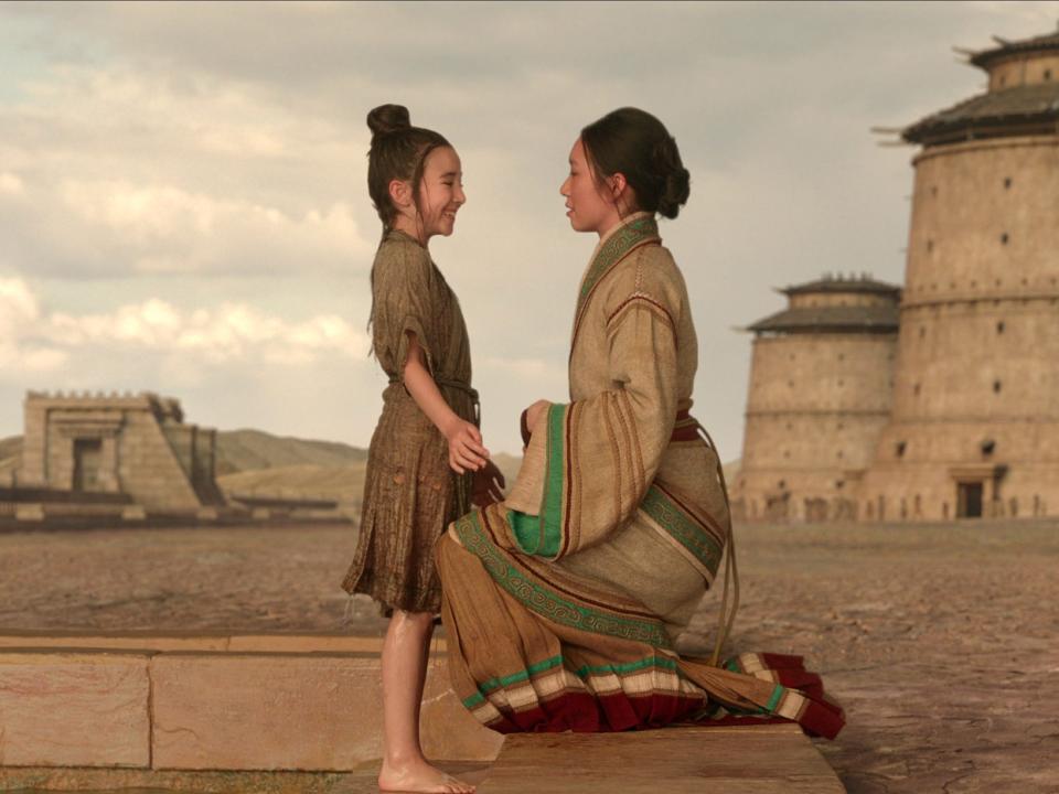 follower and jin cheng in 3 body problem. follower, a young girl, is dripping wet and smiling at jin cheng, who is kneeling on the ground to face her at eye level. in the background, there are large towers and sand