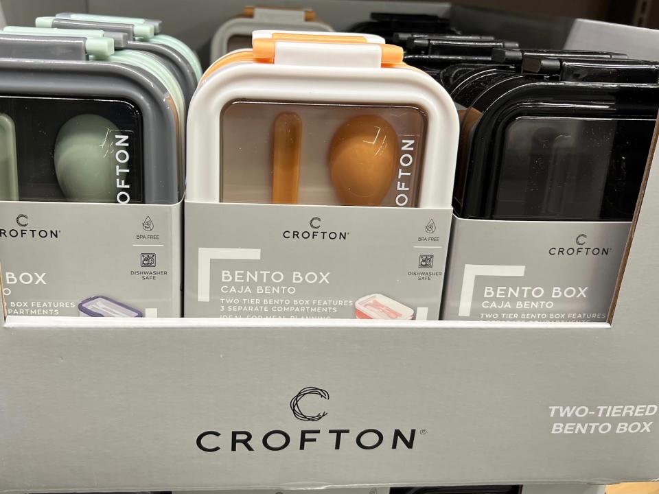 Gray cardboard boxes with Crofton labels with sets of bento boxes and silverware. The bento boxes come in green, yellow, and black colors