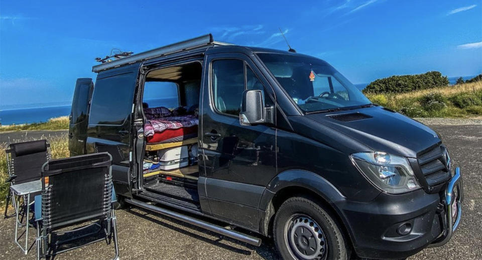 Image of a black campervan with table and chairs set up on the front of it. 