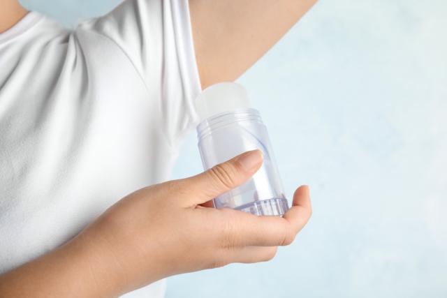 Young woman holding natural crystal alum deodorant near armpit on light blue background