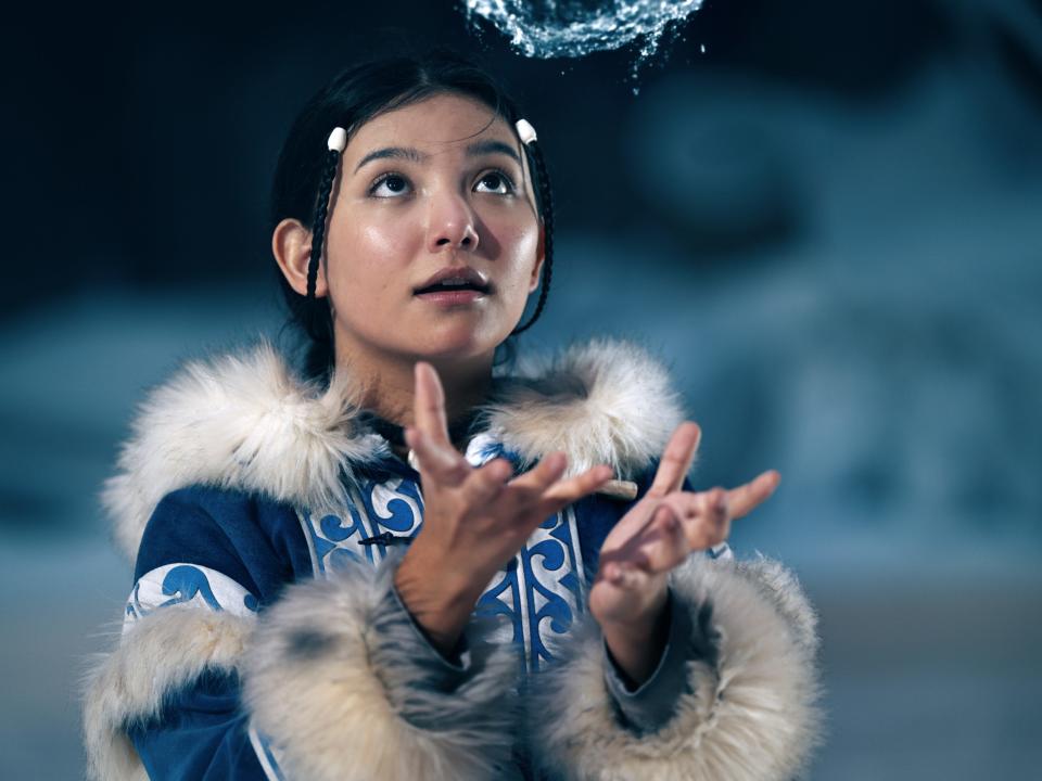 katara in the live-action netflix avatar. she's wearing blue, patterned robes with fur on the cuffs and collar, her hair pulled back with two strands looping in the front. her hands are outstretched, and she's looking at a ball of water she's making hover in the air