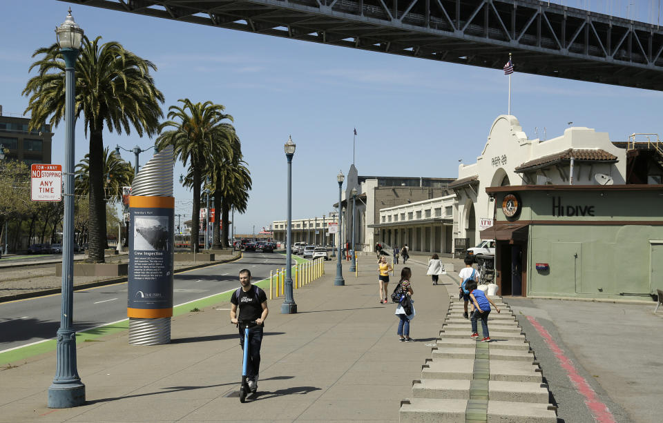 FILE - In this April 18, 2019, file photo, a man rides a scooter near children playing along the Embarcadero in San Francisco. Tired of San Francisco sidewalks being used as a testing ground for delivery robots, drones, and electric scooters, city supervisors will vote Tuesday, Dec. 10 to set up a new regulatory office that would require businesses to get permits before trying out new technology that uses public space. (AP Photo/Eric Risberg, File)