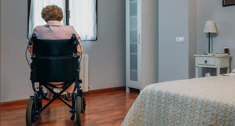 An elderly woman sits in a wheelchair in the corner of a room.