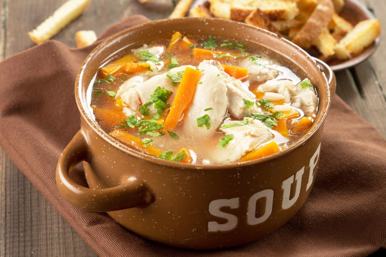 Classic chicken soup in a brown bowl with 'SOUP' etched in the side on a brown napkin with grilled cheese and fries in the background