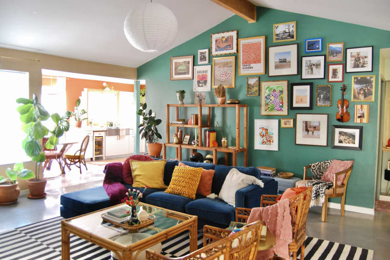 Living room with teal green gallery wall, high ceilings with wood beam, white round pendant light, black and white striped rug, blue velvet sofa with warm colored-accent pillows, cane chairs and coffee table (with glass top), wood shelves with books, art objects