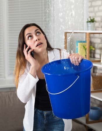 A woman makes a phone call on a cell phone while holding up a blue bucket to catch a leak.