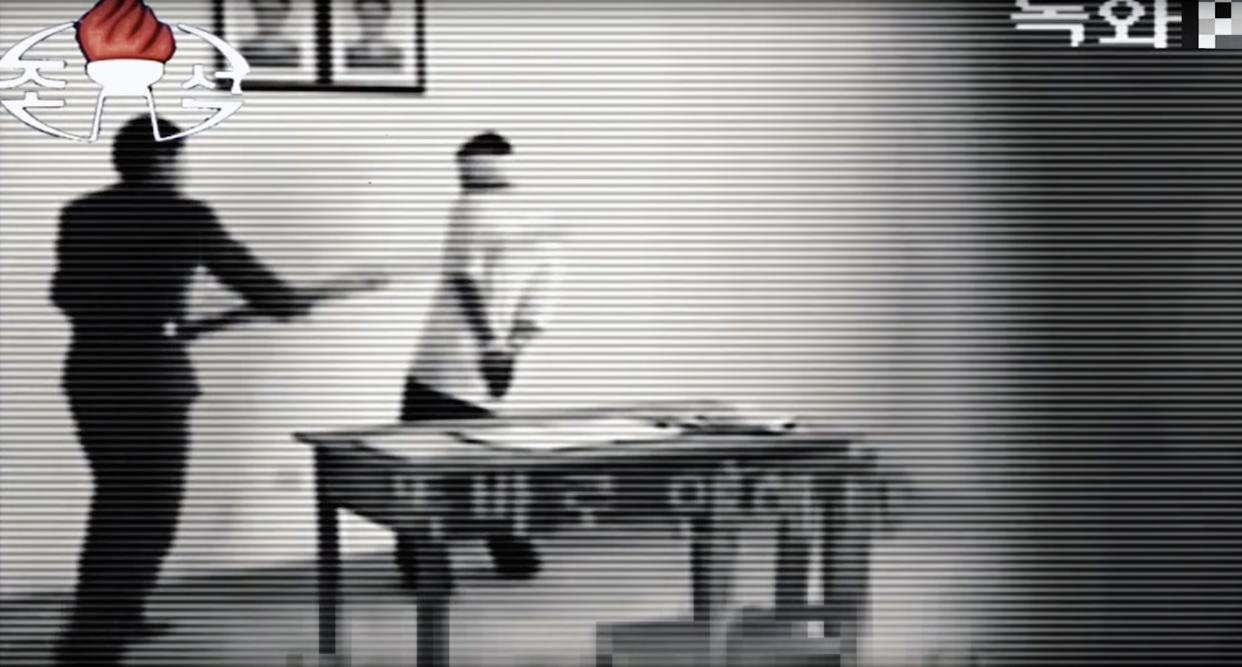 A video still of what appears to be someone beating another person with a staff in a room next to a table and below two portraits. 