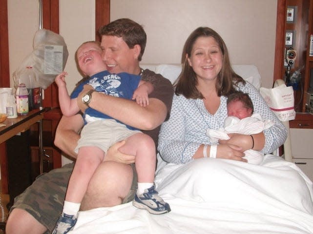 Terri Peters sitting in a hospital bed holding a newborn baby. She has dark brown hair and smiles, bleary-eyed. She wears a blue and white PJ top. Her husband sits on her right holding their son who is struggling to get out of his arms. Her husband has dark brown hair and looks to the left smiling. He wears a brown t-shirt and camo-print shorts