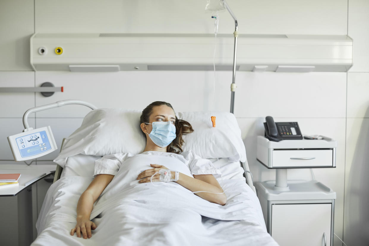 Sick female lying on bed in ICU. Mid adult patient is looking away while wearing protective face mask. She is in hospital during COVID-19 pandemic.