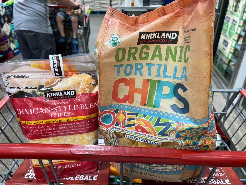 A clear plastic bag of shredded Mexican-style cheese and a brown bag of Kirkland Signature organic tortilla chips with a colorful design in a shopping cart