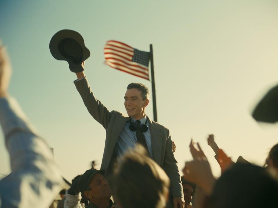 Cillian Murphy holding hat in the air as people cheer and American flag behind him