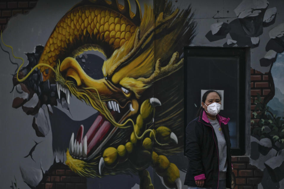 A woman wearing a face mask stands near a mural depicting a dragon in Beijing, Wednesday, Nov. 23, 2022. The latest wave of outbreaks is prompting major cities including Beijing to close off populous districts, shut stores and offices and order factories to isolate the workforces from outside contact. (AP Photo/Andy Wong)