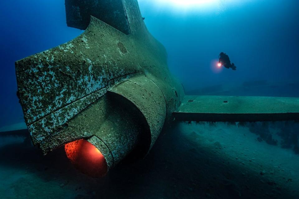 A lone diver floats near a sunken Lockheed L-1011 TriStar aircraft in Aqaba, Jordan, partially viewed from the rear. A red light placed in the engine by the photographer simulates the glow of a powered engine. The image is a winner in the 2024 Underwater Photographer of the Year Awards.
