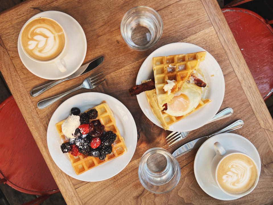 A wooden table set with two plates of waffles topped with berries and eggs, two cups of cappuccino with latte art, and glasses of water