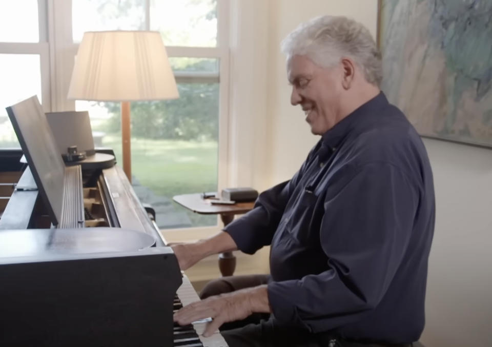 A man in a blue shirt is joyfully playing the piano in a well-lit room with a view of greenery outside