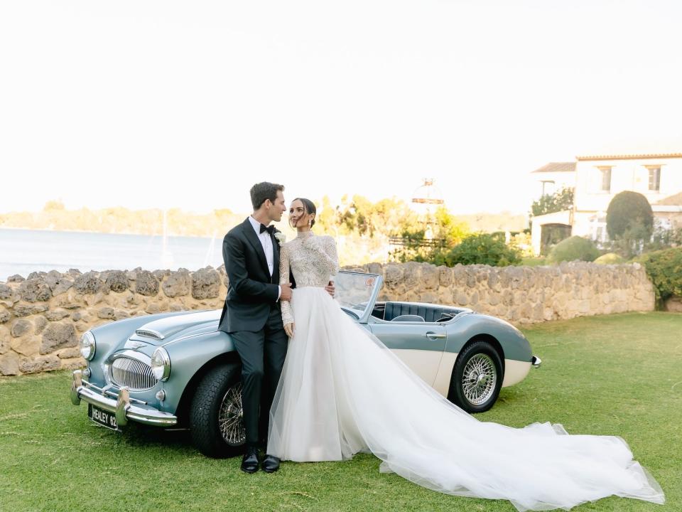 A bride and groom look at each other as they lean against a convertible.