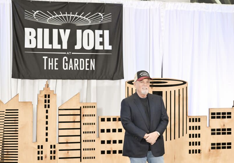 Billy Joel in a black shirt and blazer, jeans and a hat posing infant of a cloth backdrop