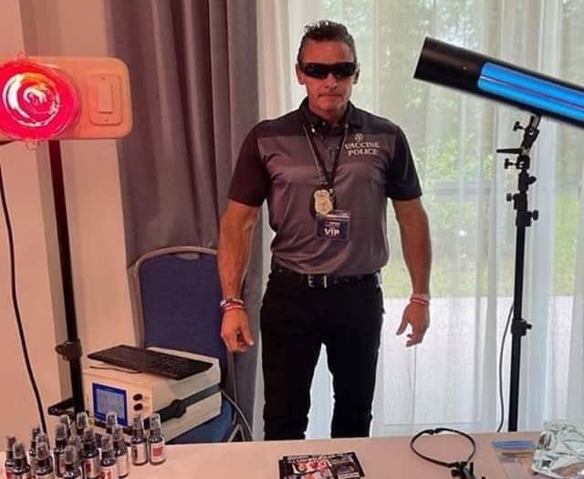 Image of a man wearing wrap around sunglasses standing rigidly behind a fold out table wearing a polo shirt with 'vaccine police' written on it. 